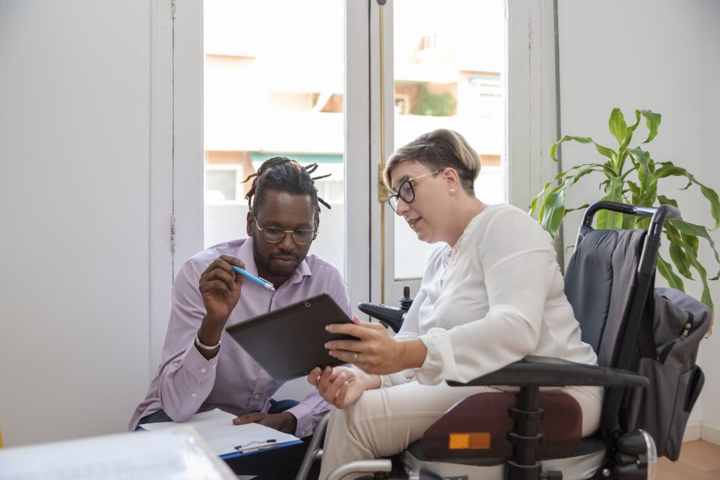 Photo image of a businesswoman in an electric wheelchair looking at a digital tablet with her male colleague who is kneeling alongside her with natural plants and window in the background