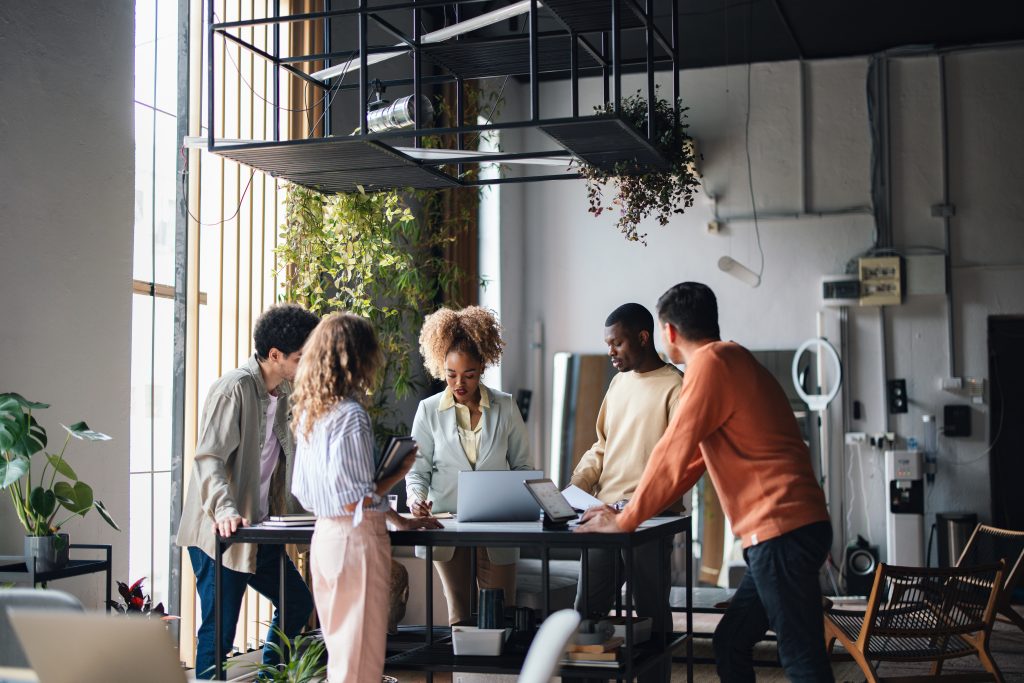 Photo image of group of diverse business people collaborating together while standing at an office desk table with laptops and tablets open in a modern industrial organic looking office setting with green plants
