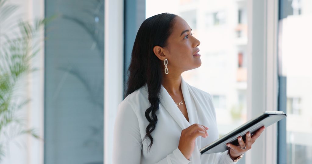 Photo image of a business woman with long black hair holding a tablet looking out a window
