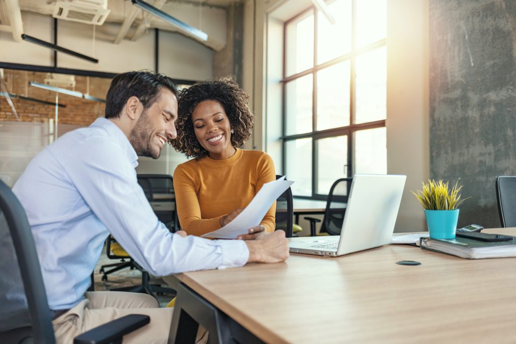 Photo image of two business people in a conference room sitting at a table with laptop open discussing the contents of a document. One is an ethnic woman and another is a caucasian male with a modern background and window